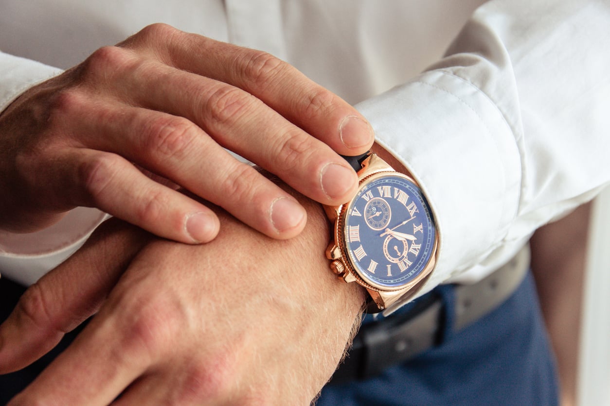A person in a white dress shirt adjusts a gold wristwatch with a blue face and Roman numerals on their left wrist—one of those classic products men skip buying but eventually realize they need.