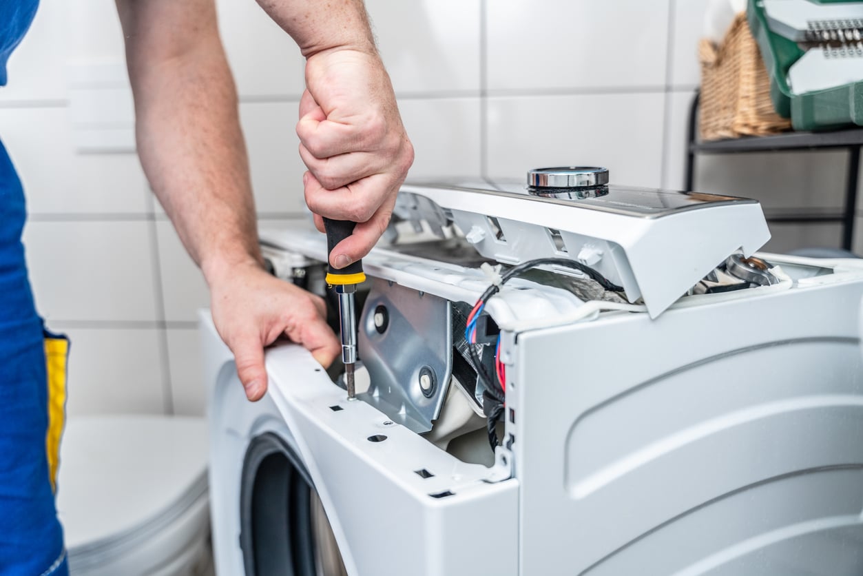 A person uses a screwdriver to repair the top panel of a washing machine, with the machine partially disassembled in a tiled laundry room.