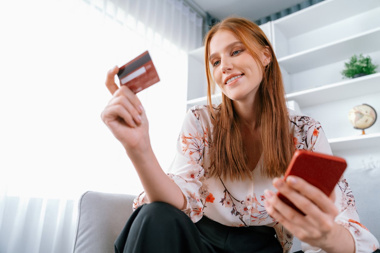 A young woman with long red hair smiles while holding a credit card in one hand and a smartphone in the other, sitting on a couch in a bright, modern living room.