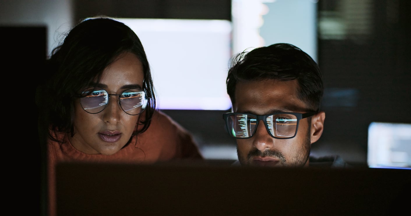 Two people in glasses closely look at computer screens in a dimly lit room, with code reflected in their lenses, suggesting they are working together on a programming or technical task.