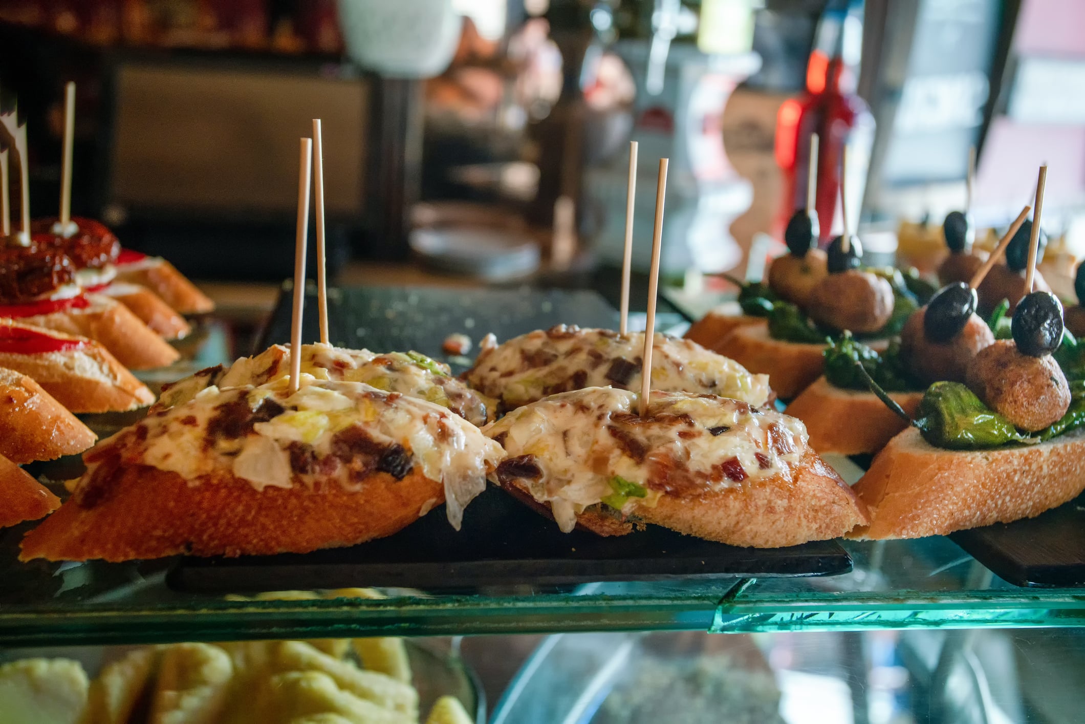 Slices of bread topped with creamy spreads, meat, and vegetables are displayed on a glass shelf, each piece held together with wooden toothpicks. The background is blurred, highlighting the colorful appetizers.
