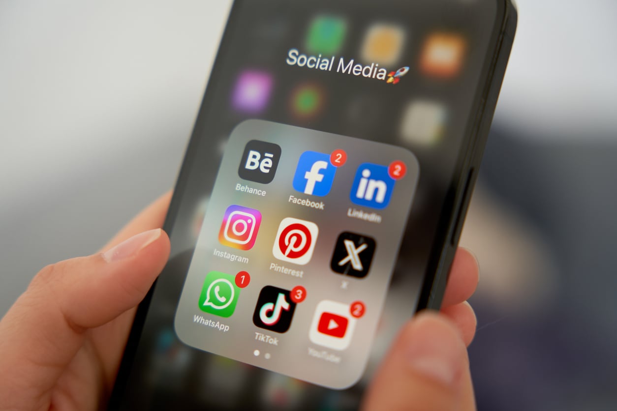 A person holds a smartphone displaying a folder of social media apps, including Behance, Facebook, LinkedIn, Instagram, Pinterest, WhatsApp, TikTok, and YouTube—some with notification alerts—highlighting the impact of notifications on mental health.