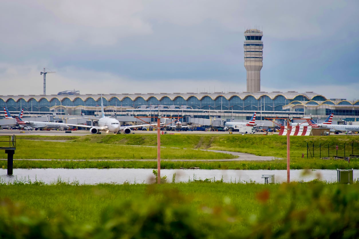 A busy airport scene shows multiple airplanes parked at gates in front of a terminal, highlighting one of the most convenient airports, with an air traffic control tower, grassy areas, and water in the foreground beneath a cloudy sky.