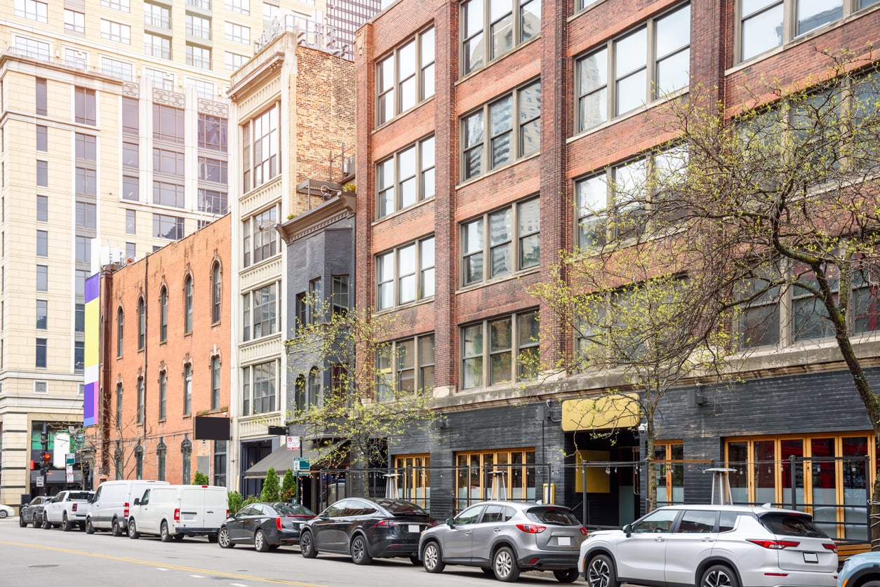 A city street lined with parked cars and various multi-story brick and glass buildings, featuring shops and trees with sparse leaves along the sidewalk—a vibrant scene common in thriving Small Business States.