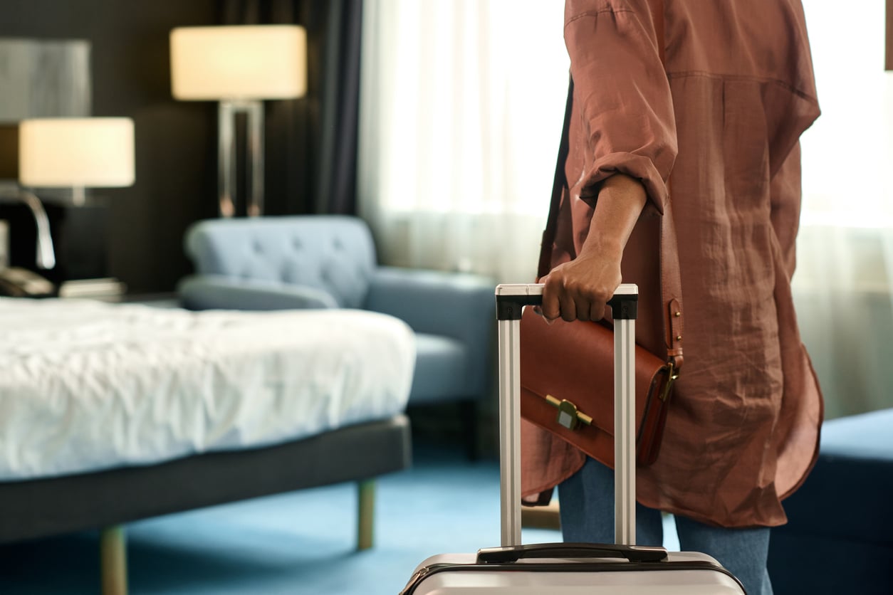 A person stands in a hotel room, holding the handle of a suitcase. Emphasizing hotel room safety, the space features a made bed, a blue armchair, and large windows letting in natural light.
