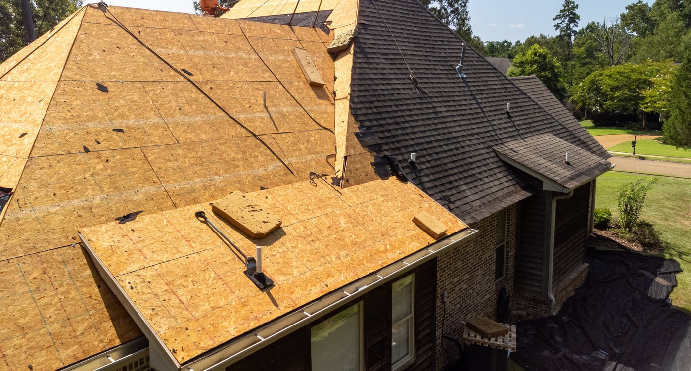 A house roof under repair, with part of the roof covered in wooden sheathing and part still covered with shingles—tools and materials from delayed purchases are visible on the roof, with a yard and trees in the background.