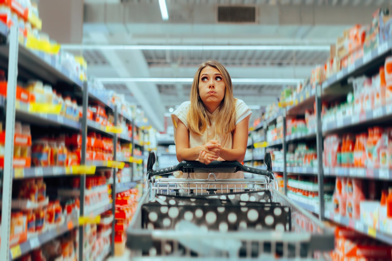 A woman leans on a shopping cart in a grocery store aisle, looking up with a thoughtful, overwhelmed expression as she considers the many overpriced products lining the colorful shelves around her.