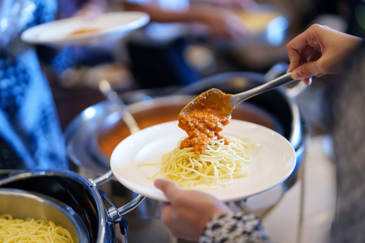 A person serves tomato-based sauce onto a plate of plain spaghetti noodles at a buffet, with more food trays visible in the background.