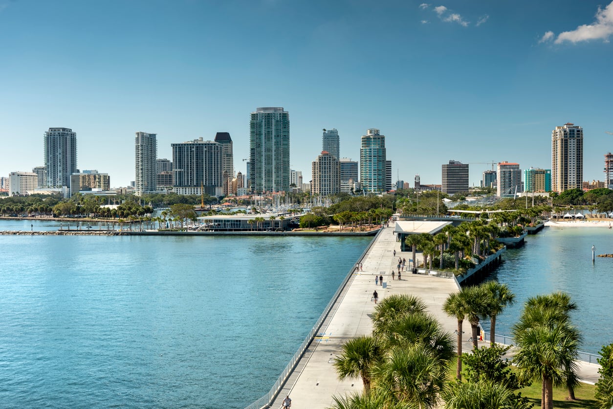 View of a waterfront city skyline with tall buildings, a pier extending into the water, palm trees, and people walking along the pier under a clear blue sky.