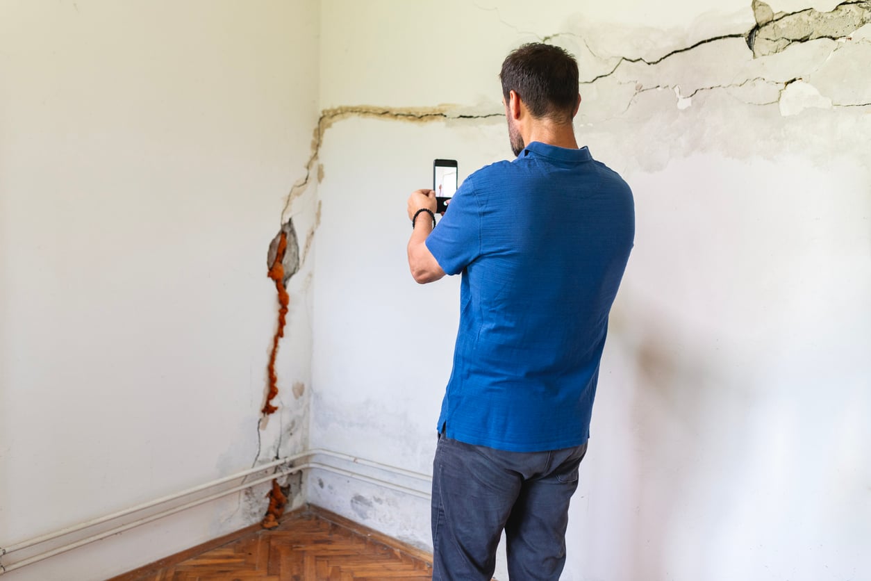 A man in a blue shirt, staying at an Airbnb, takes a photo with his phone of a large crack and water damage on a white wall in the corner of a room with wooden floors.