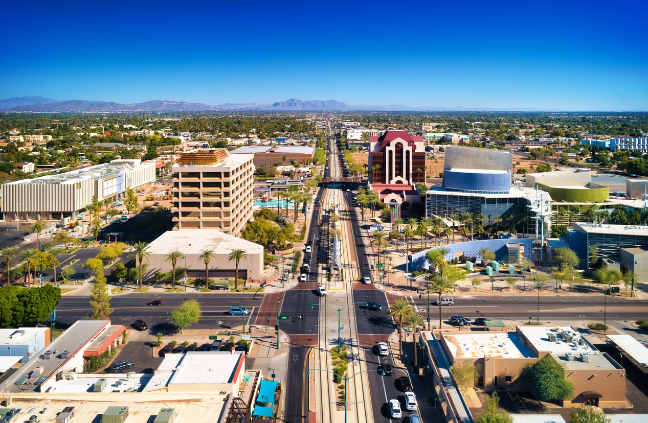 Aerial view of a cityscape with palm-lined streets, modern buildings, cars, and distant mountains under a clear blue sky.