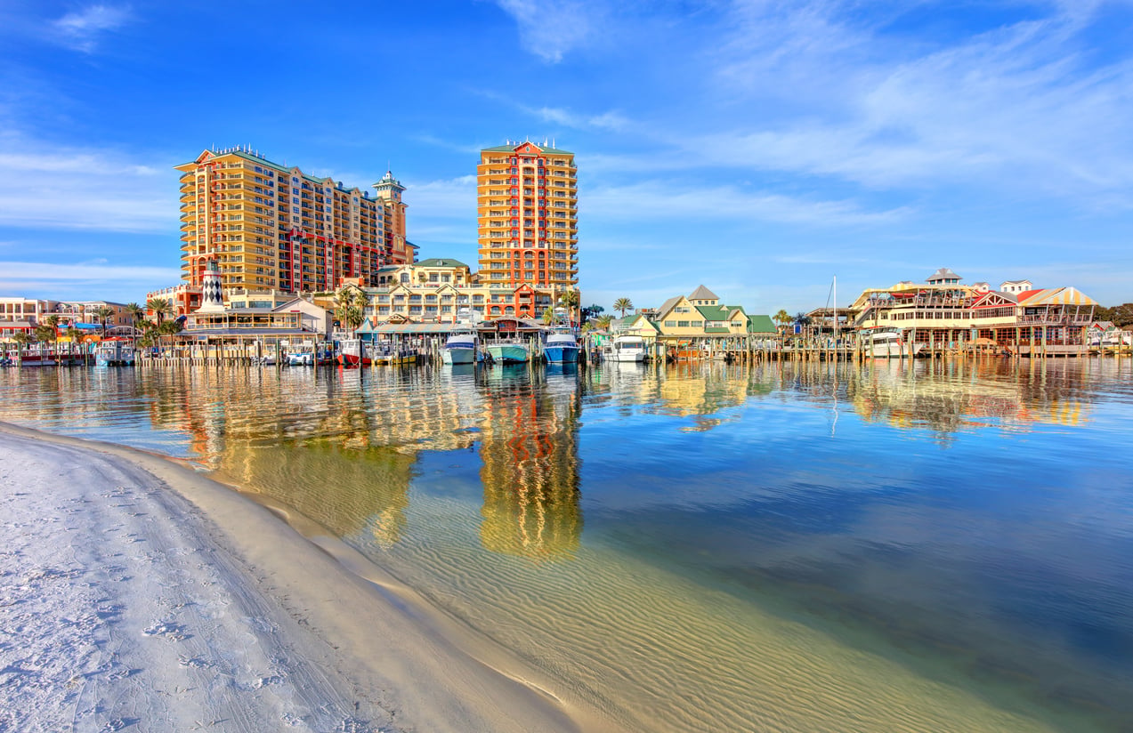 Colorful beachfront buildings and boats are reflected in calm water under a blue sky, with a sandy shoreline in the foreground.