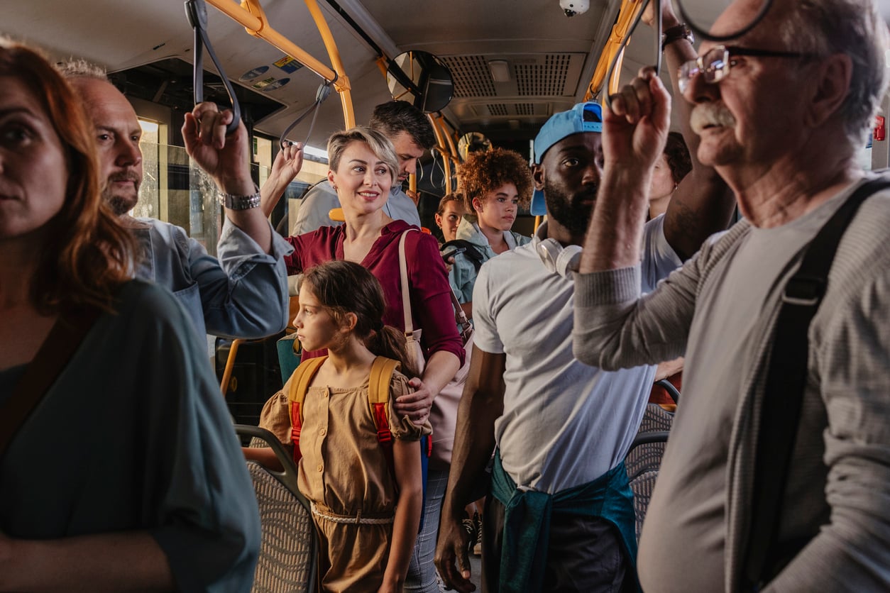 A diverse group of people stand closely together on a crowded city bus, some holding onto handrails. A woman in red smiles, a young girl with a backpack looks to the side, and others appear thoughtful or focused.