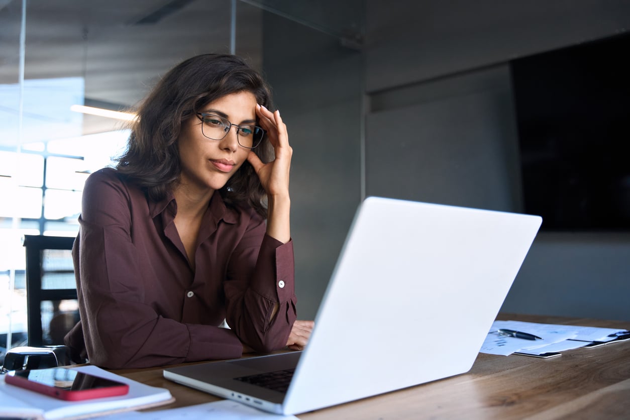 A businesswoman with glasses rests her forehead on her hand while looking at a laptop in an office.