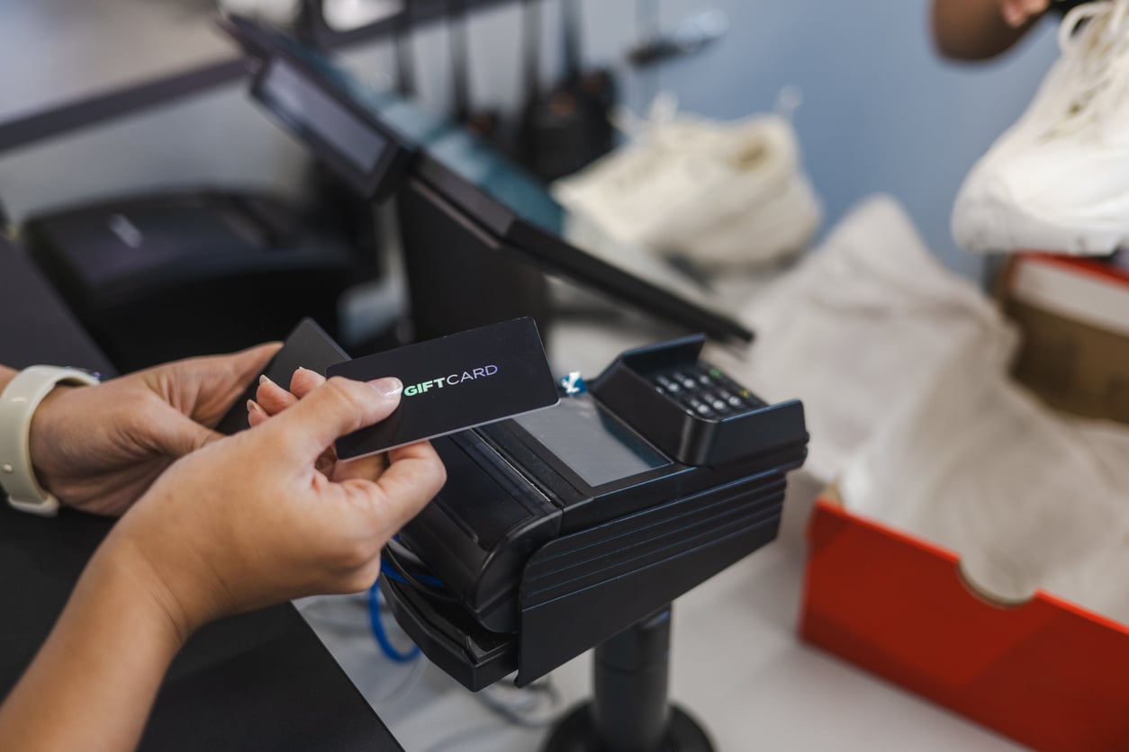 A person holds a gift card near a payment terminal at a store counter, preparing to make a purchase. Sneakers and a shoebox are visible in the background—perfect for those seeking travel hacks to save on essential gear.