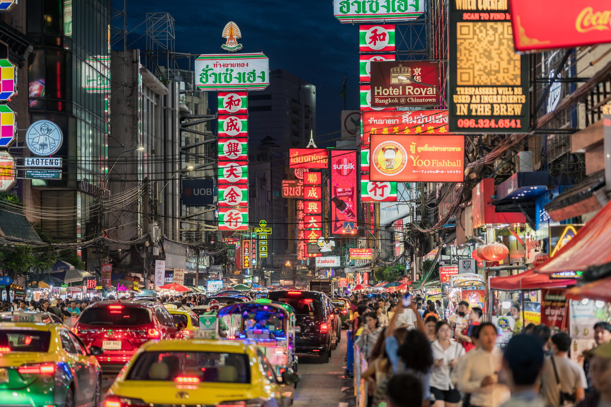A busy street at night in an urban area, filled with cars, people, and bright neon signs with Thai and Chinese characters, creating a vibrant and lively atmosphere.