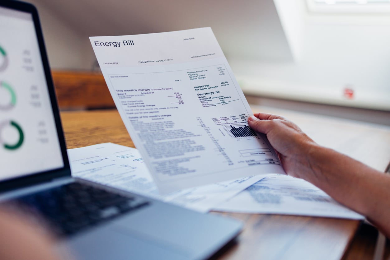 A person holds an energy bill next to a laptop, with other documents spread on the table, suggesting they are reviewing household expenses or budgeting.