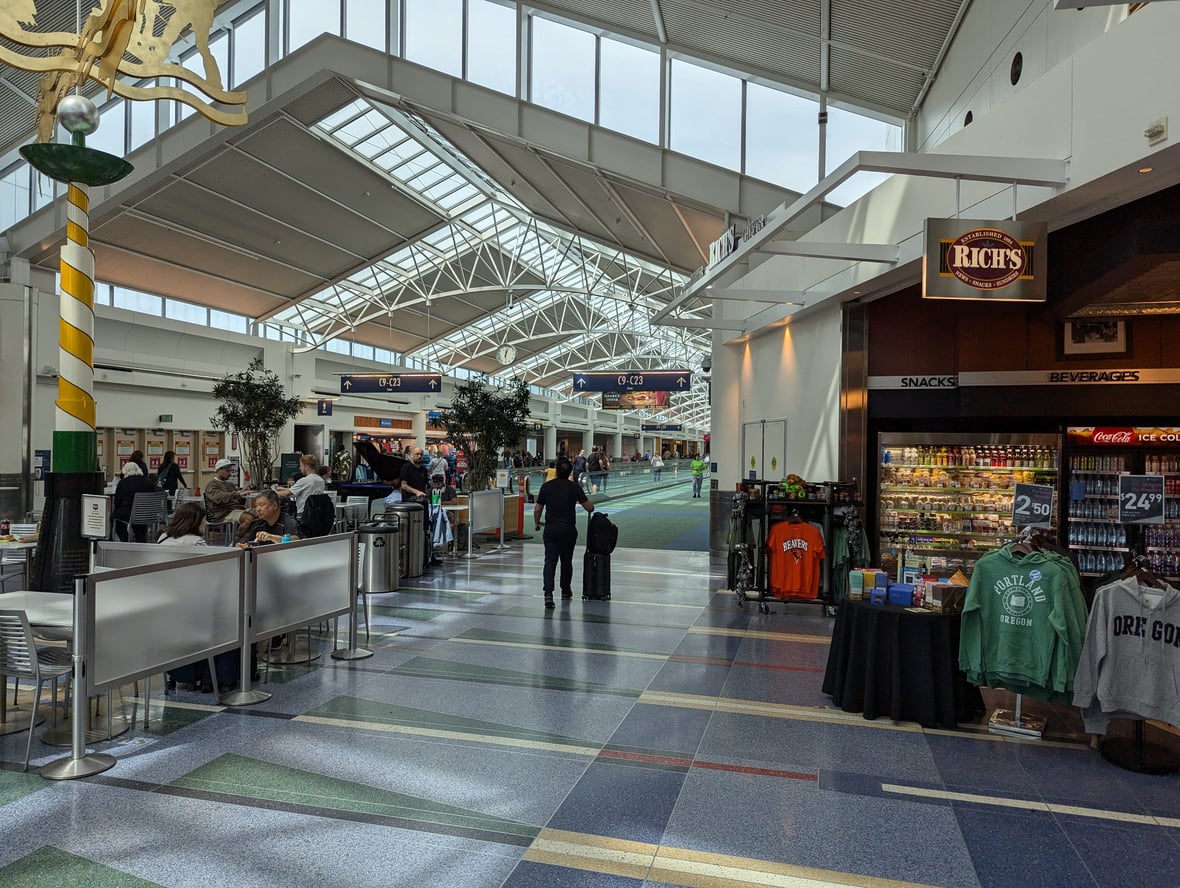 A busy airport terminal with people sitting and walking, some with luggage. A snack shop with drinks and Oregon-themed merchandise is on the right. Large windows and skylights provide natural light in one of the most convenient airports. Signs for gates and shops are visible.