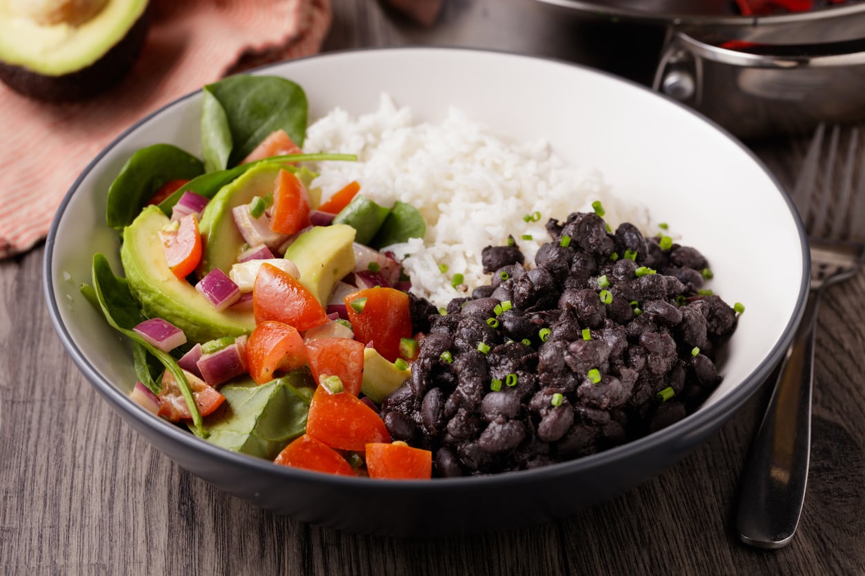 A bowl filled with white rice, seasoned black beans—a great ground beef alternative—sliced avocado, spinach, and a colorful salad of tomatoes and red onions, set on a wooden table with a fork and avocado in the background.