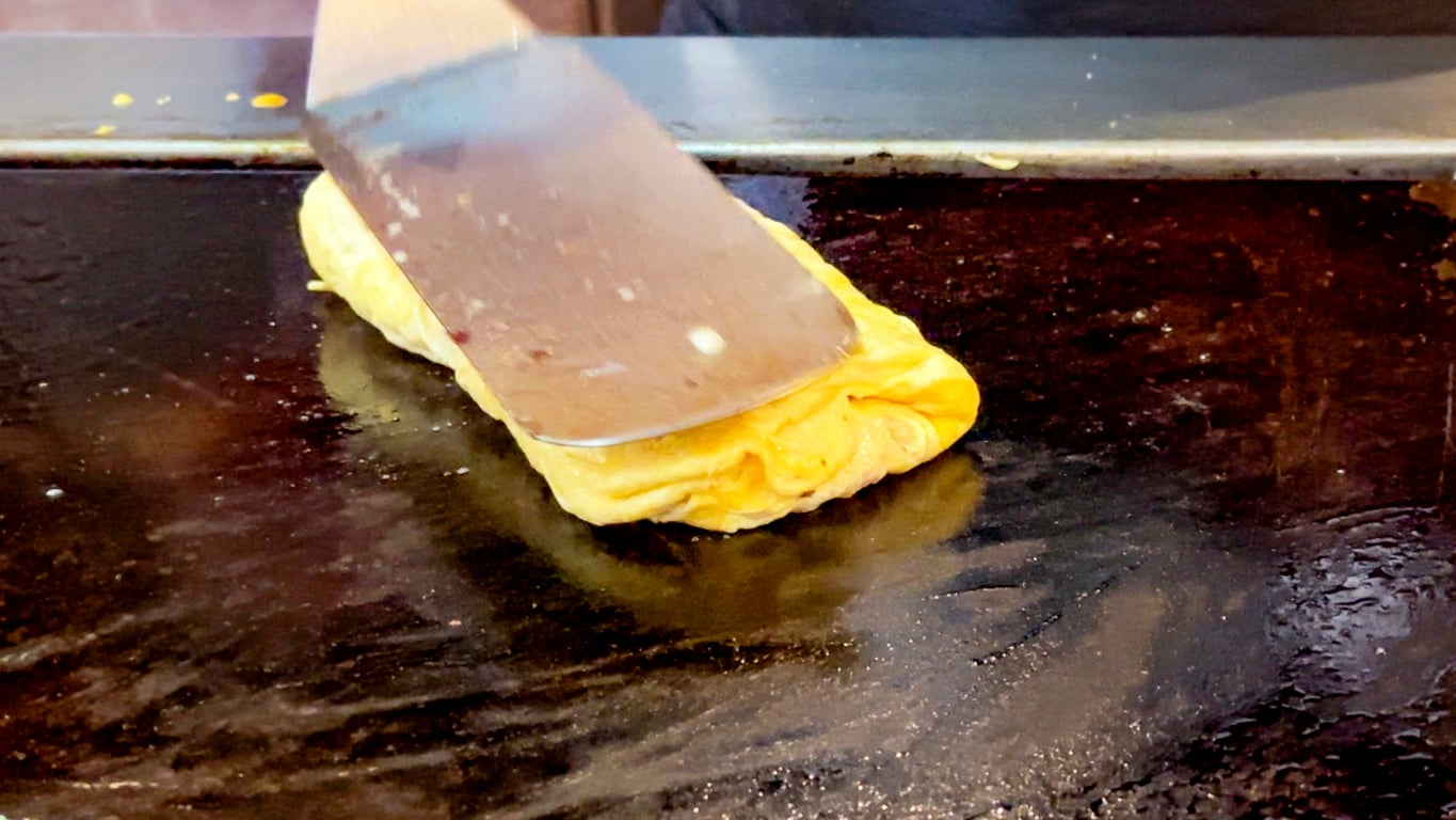 A close-up of a metal spatula folding a rectangular yellow omelette on a dark, oily griddle.