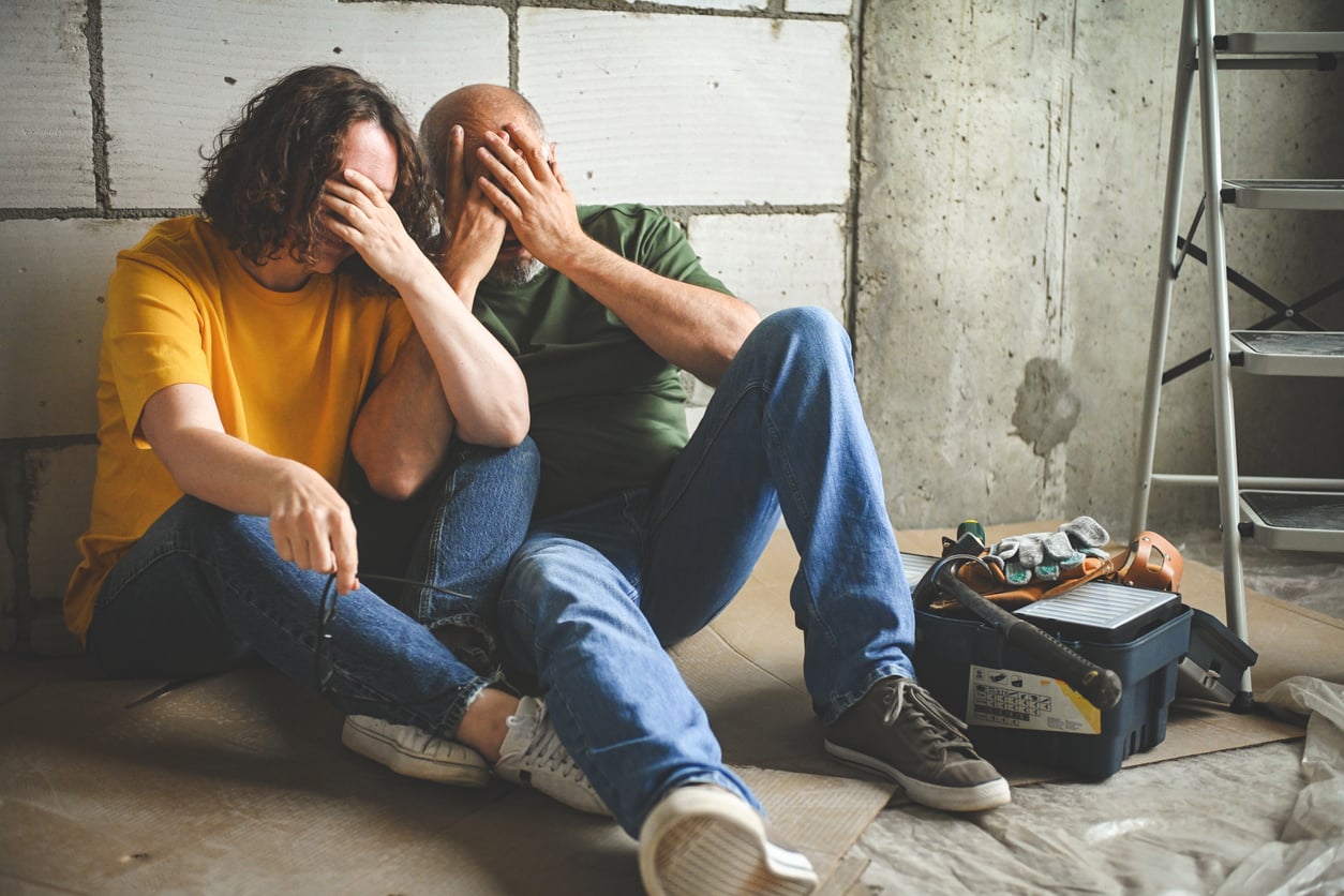 Two people sit on the floor of a room under renovation, looking frustrated with their hands covering their faces. A toolbox and tools are next to them, and a ladder is nearby against a concrete wall.