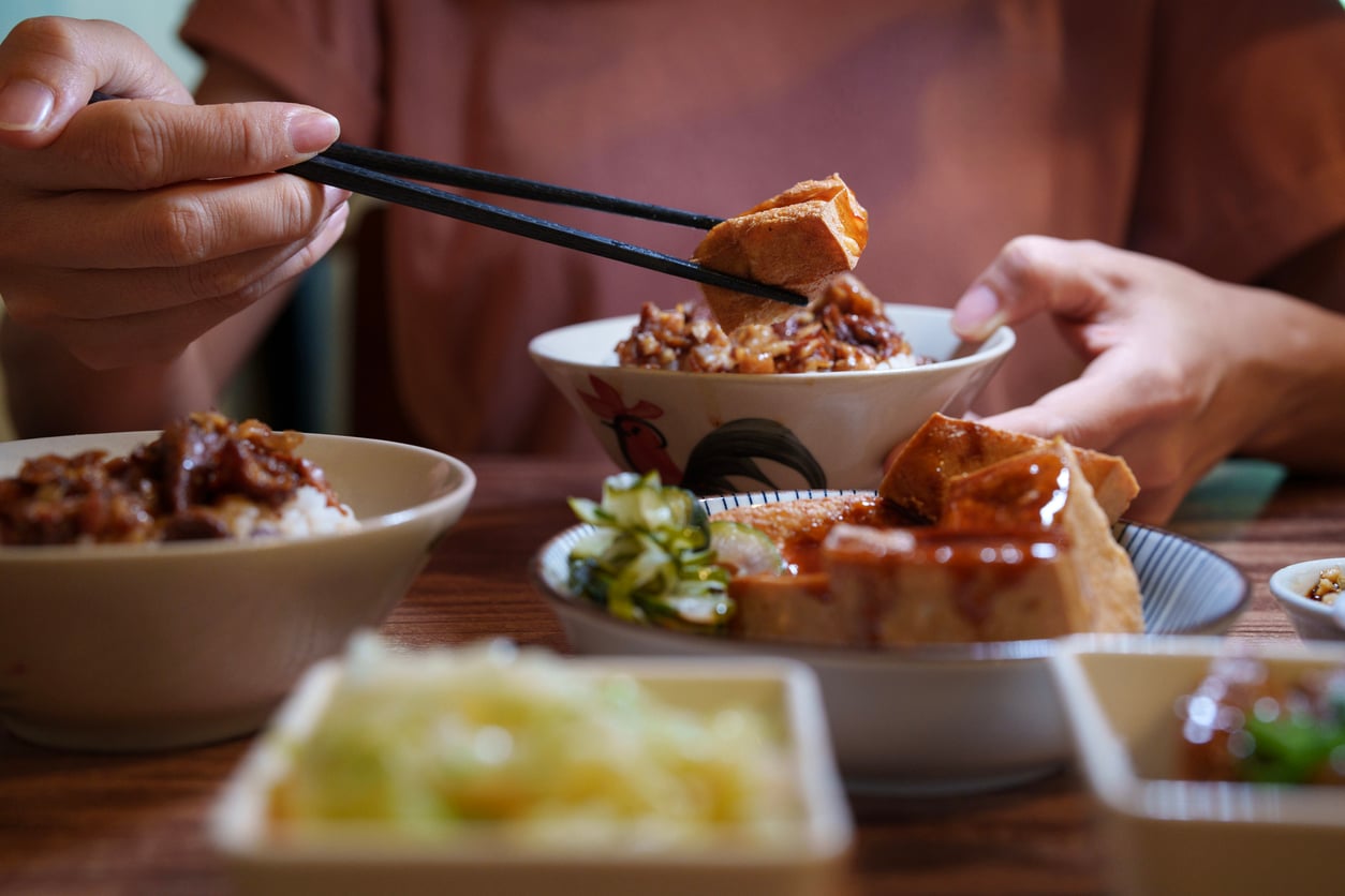 A person uses chopsticks to pick up a piece of food from a bowl, with several bowls and dishes of rice, vegetables, tofu, and other ground beef alternatives on a wooden table in front of them.