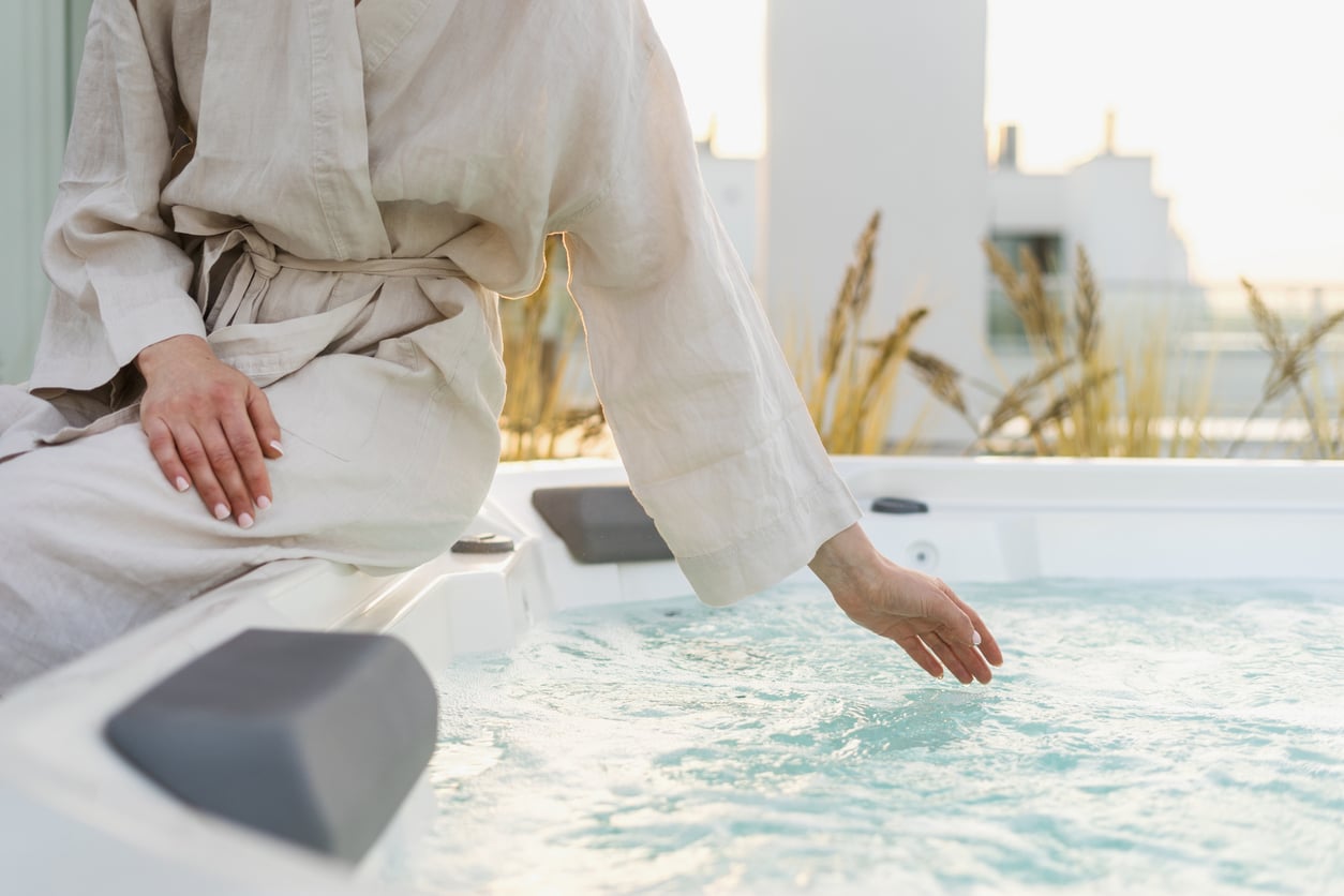 A person wearing a beige robe sits on the edge of a hot tub, gently touching the bubbling water with their hand. Grassy plants and sunlight are visible in the background.