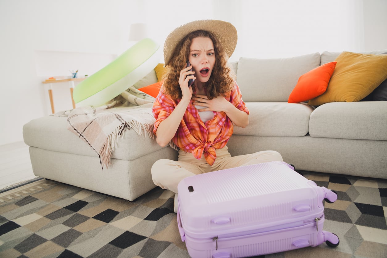 A surprised woman in a sun hat and plaid shirt sits on the floor by a lavender suitcase, talking on her phone in a bright Airbnb living room with colorful cushions and an inflatable pool float.