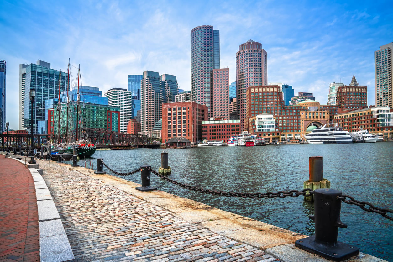 Boston waterfront with a view of modern and historic skyscrapers, boats docked in the harbor, and a cobblestone walkway lined with a metal chain fence under a blue sky—far from worries like credit card debt.