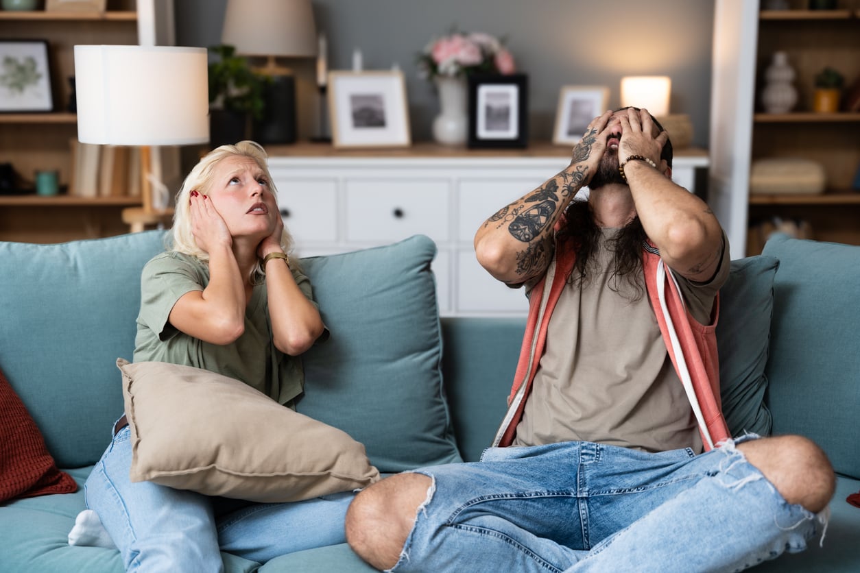 Two people sitting on a couch appear frustrated; one covers their ears and looks up, while the other covers their face with both hands. The room is decorated with shelves and framed pictures in the background.