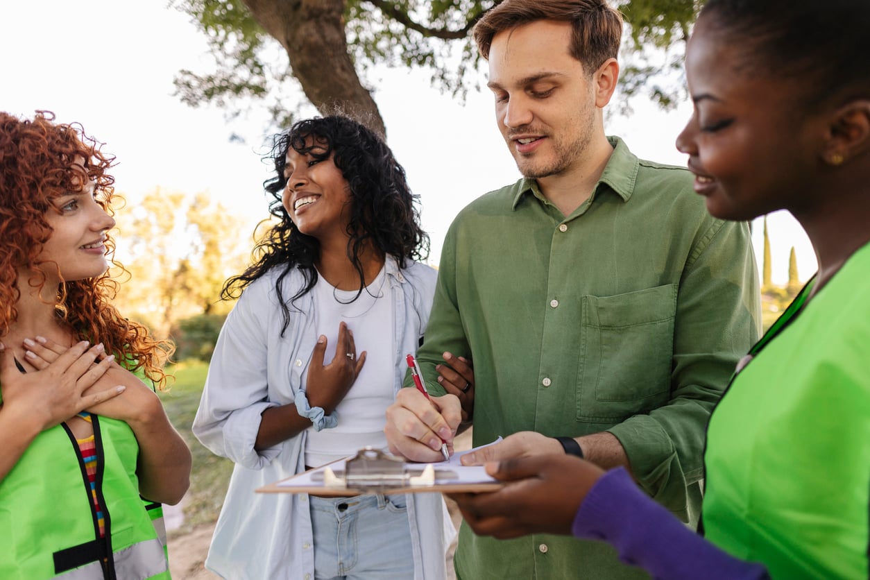 Four people stand outdoors, smiling and interacting as they discuss budget-friendly hobbies. One person holds a clipboard while another signs it. The group appears cheerful and engaged, with trees and sunlight in the background.