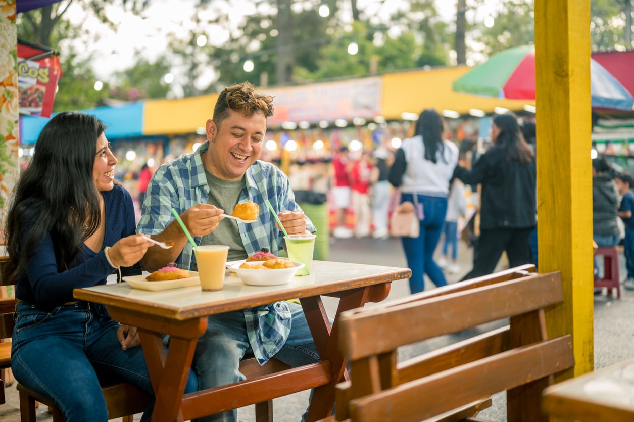 Two people sit at an outdoor table enjoying food and drinks, smiling and laughing together while sharing theme park hacks. The background shows a busy fair or market with colorful booths, people walking, and trees.