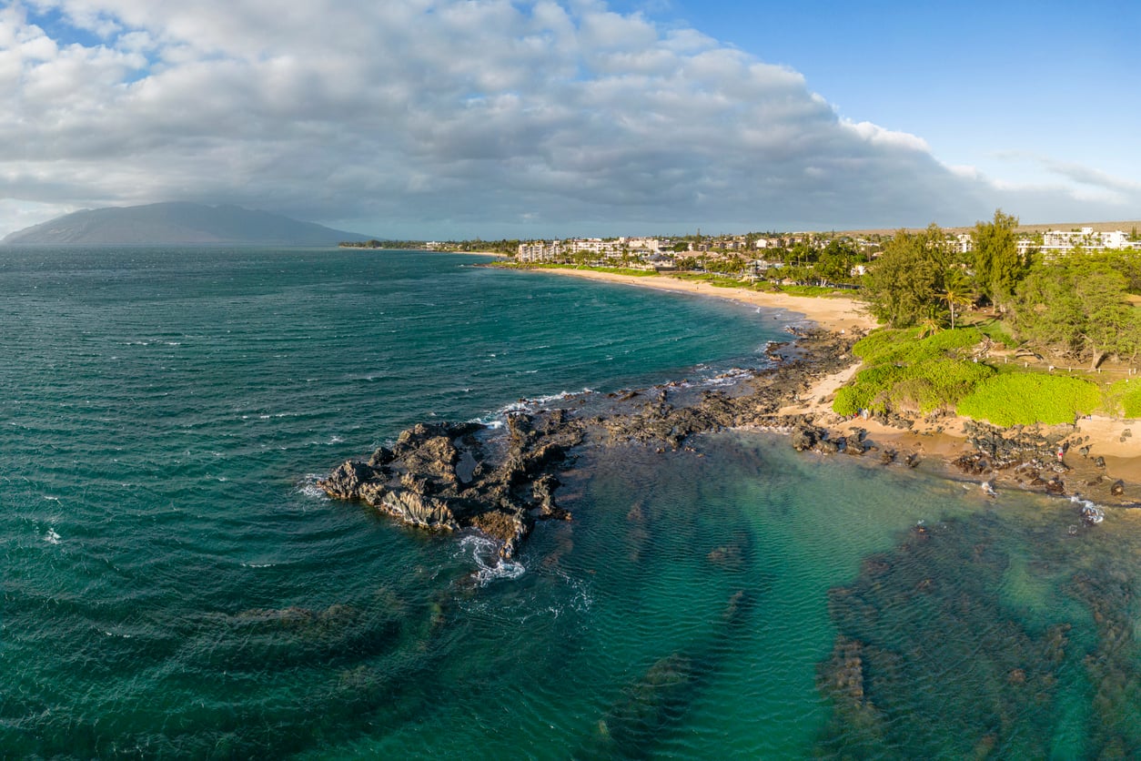Aerial view of a scenic coastline with turquoise ocean, rocky outcrops, sandy beach, and lush greenery, with buildings and mountains in the background under a partly cloudy sky.