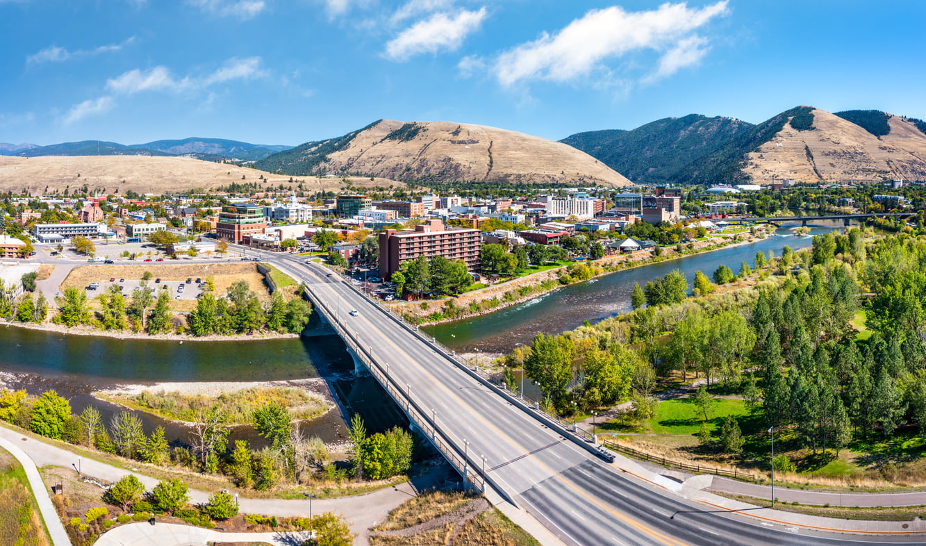 A wide aerial view of a bridge crossing a river into an affordable college town surrounded by green trees, with tan hills and blue mountains in the background under a bright, partly cloudy sky.