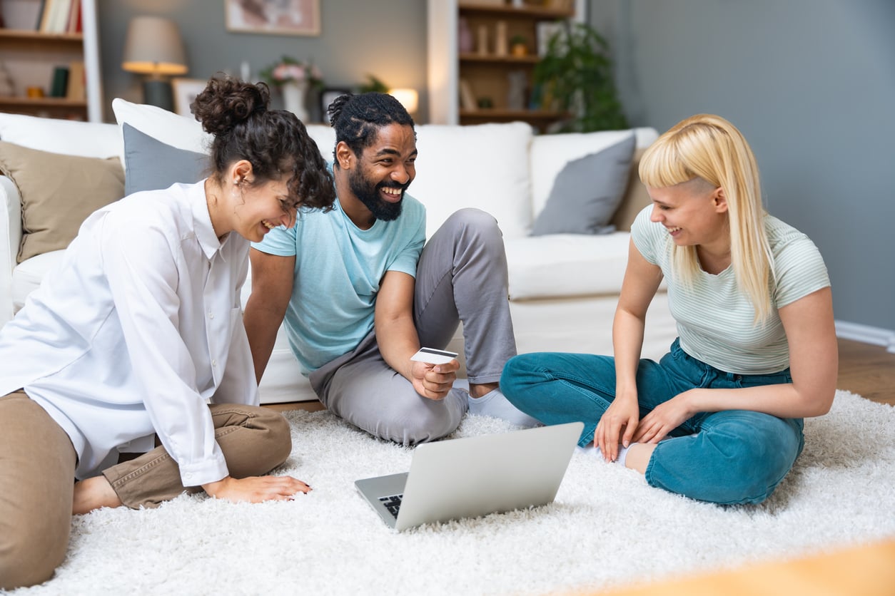 Three people sit on a carpeted floor, smiling and looking at a laptop together. One person holds a credit card, suggesting they are shopping or making a payment online in a cozy living room setting.