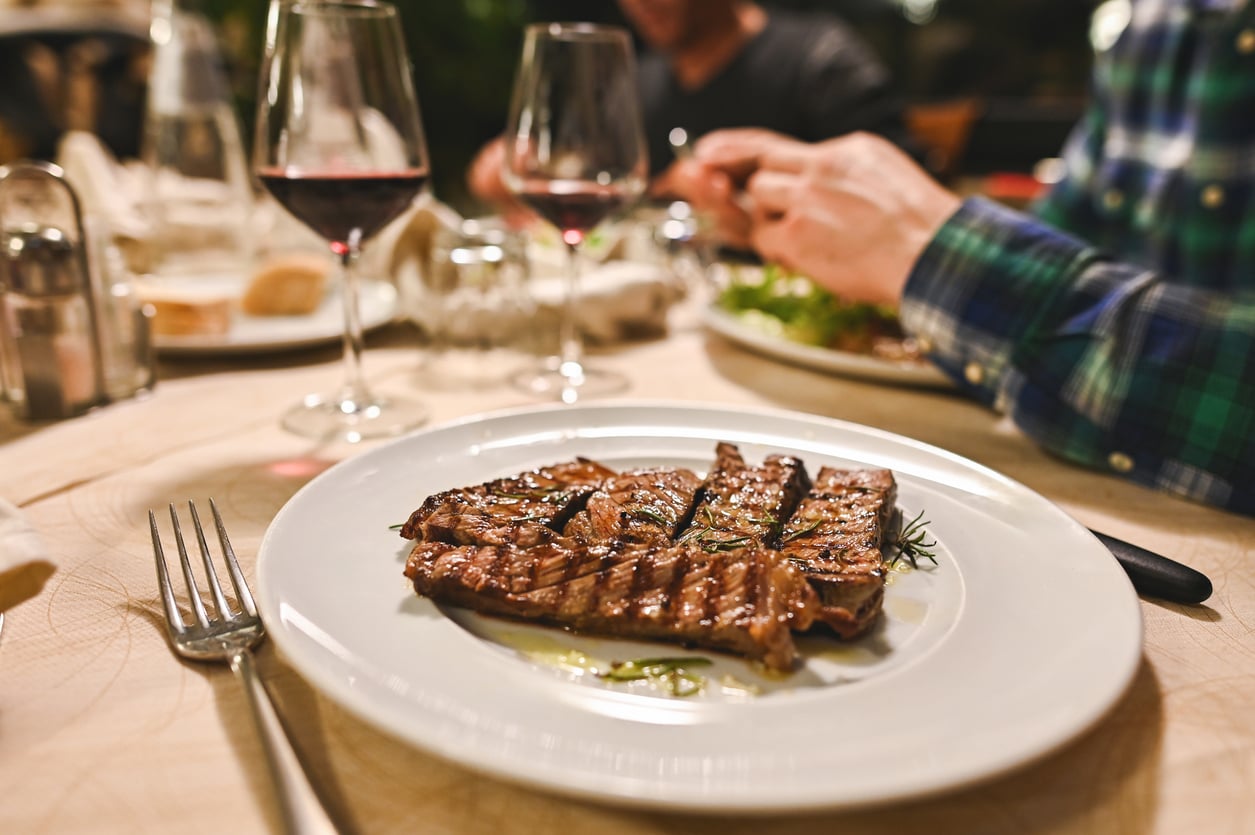 A grilled steak garnished with rosemary is served on a white plate, with two glasses of red wine—an indulgence often found among products men skip buying—and a person dining in the background at a restaurant table.