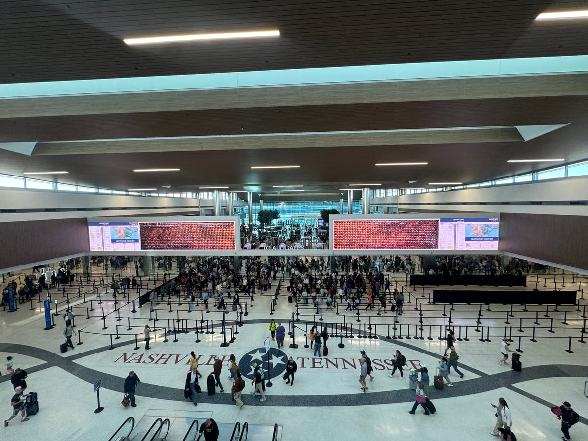 A busy airport terminal in Nashville, Tennessee, known as one of the most convenient airports, with large crowds in security lines, digital flight information screens, and a star emblem on the floor reading "Nashville Tennessee.