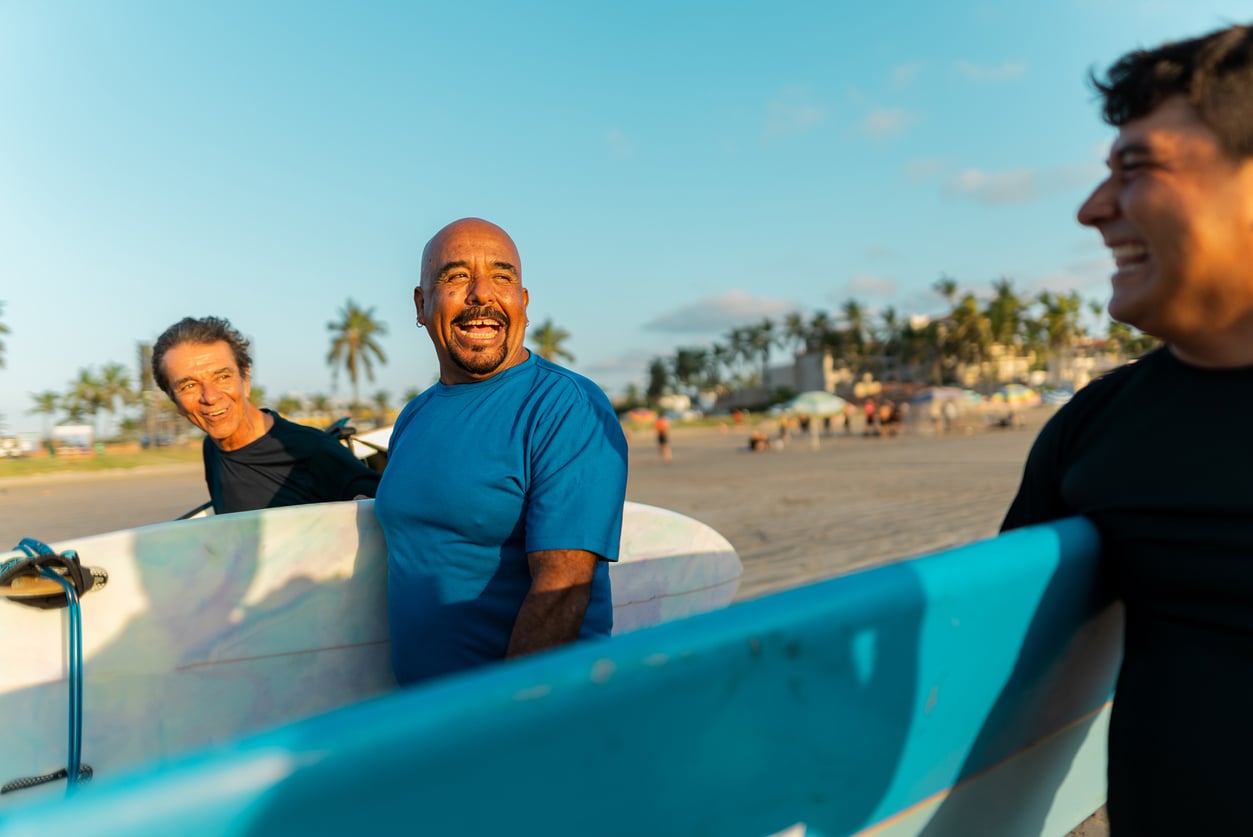 Three men in wetsuits smile and carry surfboards on a sunny beach, with palm trees and people in the background.