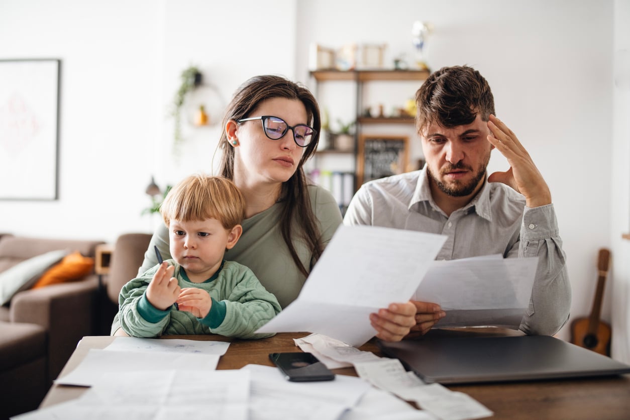 A worried family of three, with parents looking at bills and a young child sitting between them, sits at a table covered in papers in a home setting.