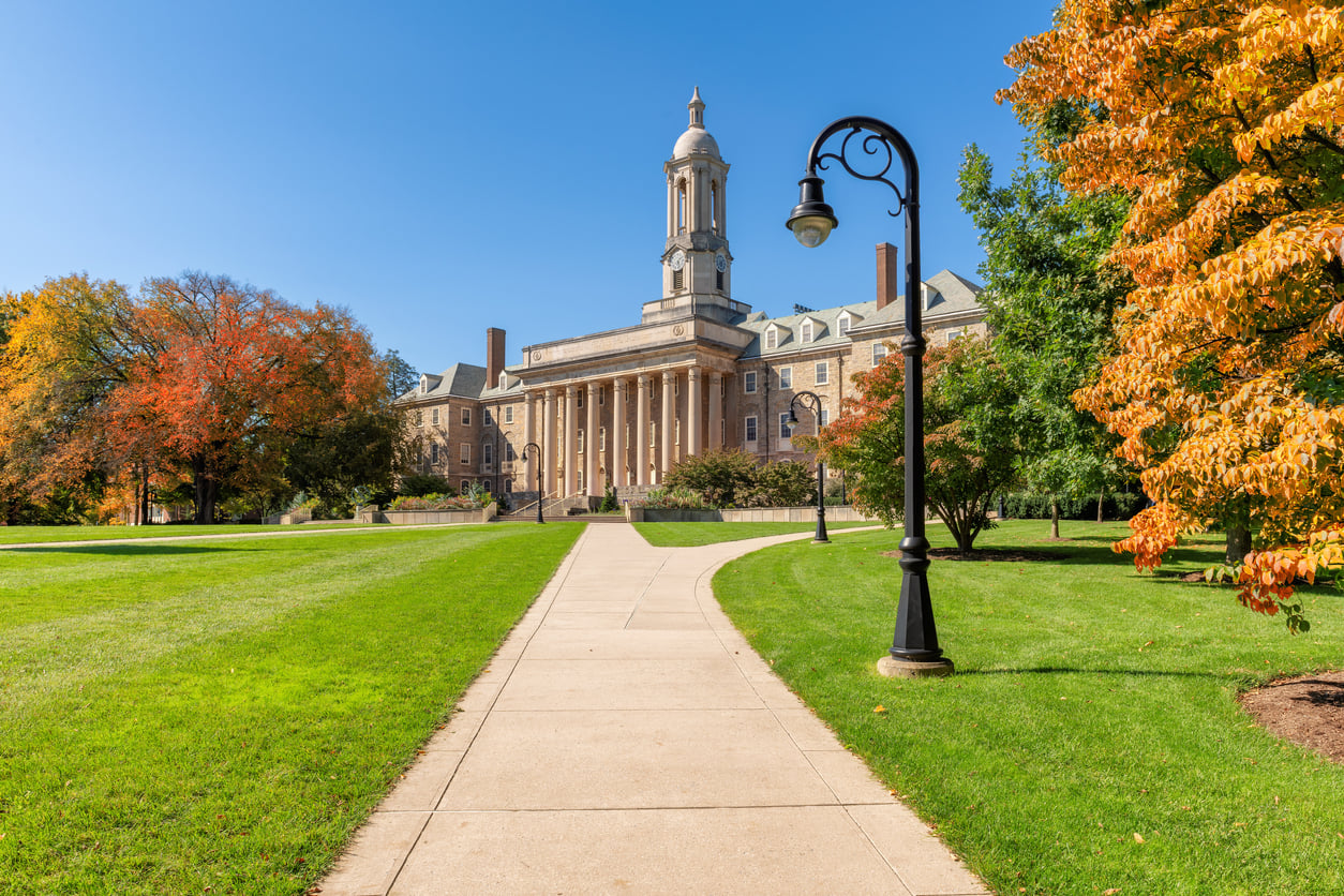 A wide sidewalk leads to a large, historic building with columns and a clock tower, nestled in one of the most affordable college towns, surrounded by green lawns and trees with autumn foliage under a clear blue sky.