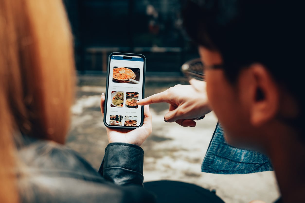 Two people sitting together, with one holding a smartphone displaying images of food. The other person is pointing at the screen, suggesting they are choosing or discussing food options, possibly for ordering.