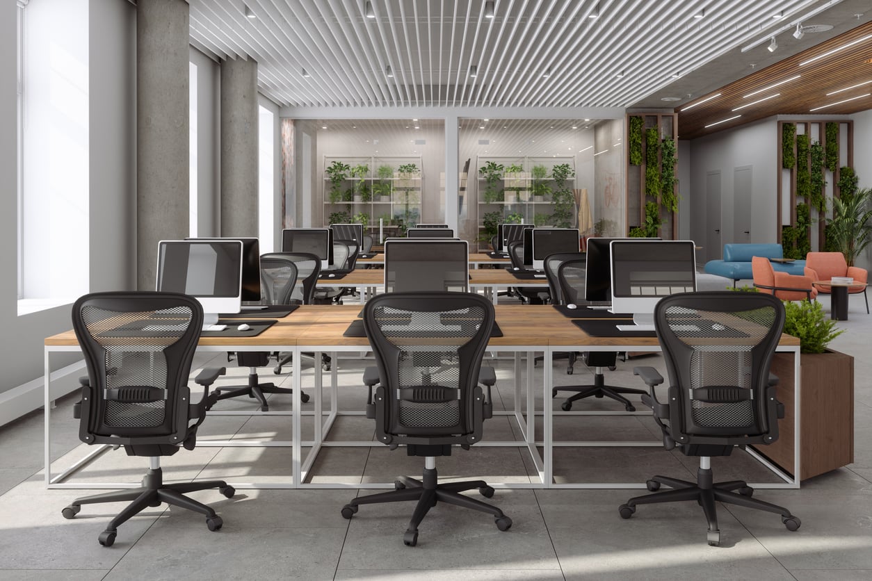 Modern open-plan office with rows of empty desks, ergonomic chairs, and computer monitors. Large windows, indoor plants, and natural light create a bright, spacious, and inviting workspace.