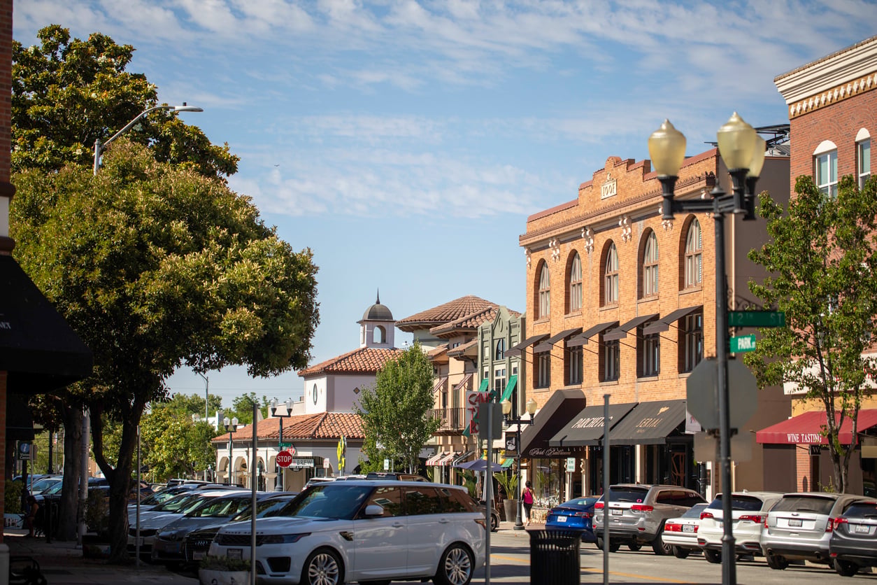 Cars are parked along a sunny street lined with trees and historic brick buildings, with shops and cafes visible. A small white clock tower rises in the background under a partly cloudy blue sky.