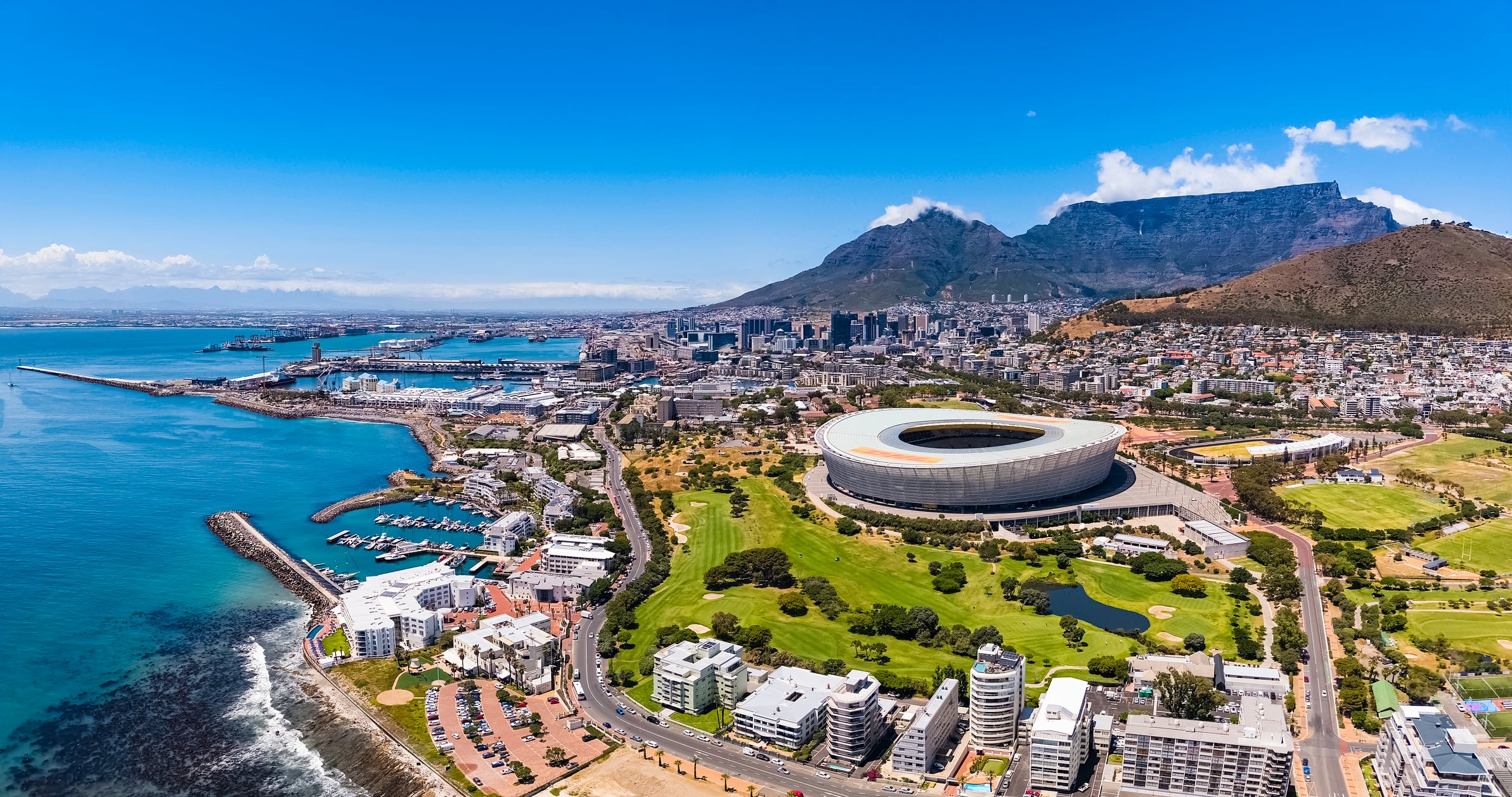 Aerial view of Cape Town, South Africa, featuring the Cape Town Stadium near the coast, green spaces, city buildings, and Table Mountain in the background under a clear blue sky.