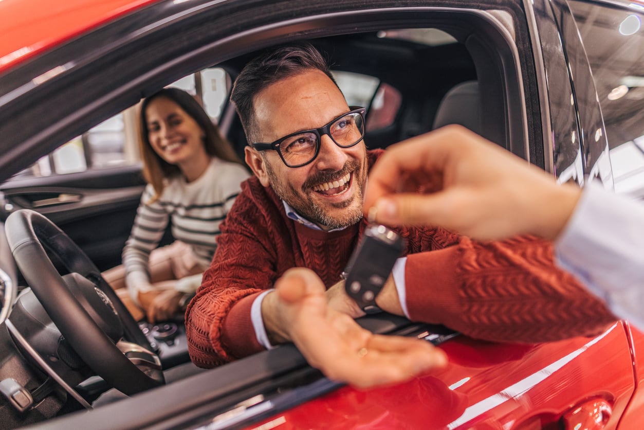 A smiling man sitting in the driver’s seat of a red car reaches for car keys from someone outside, while a woman in the passenger seat looks on happily.