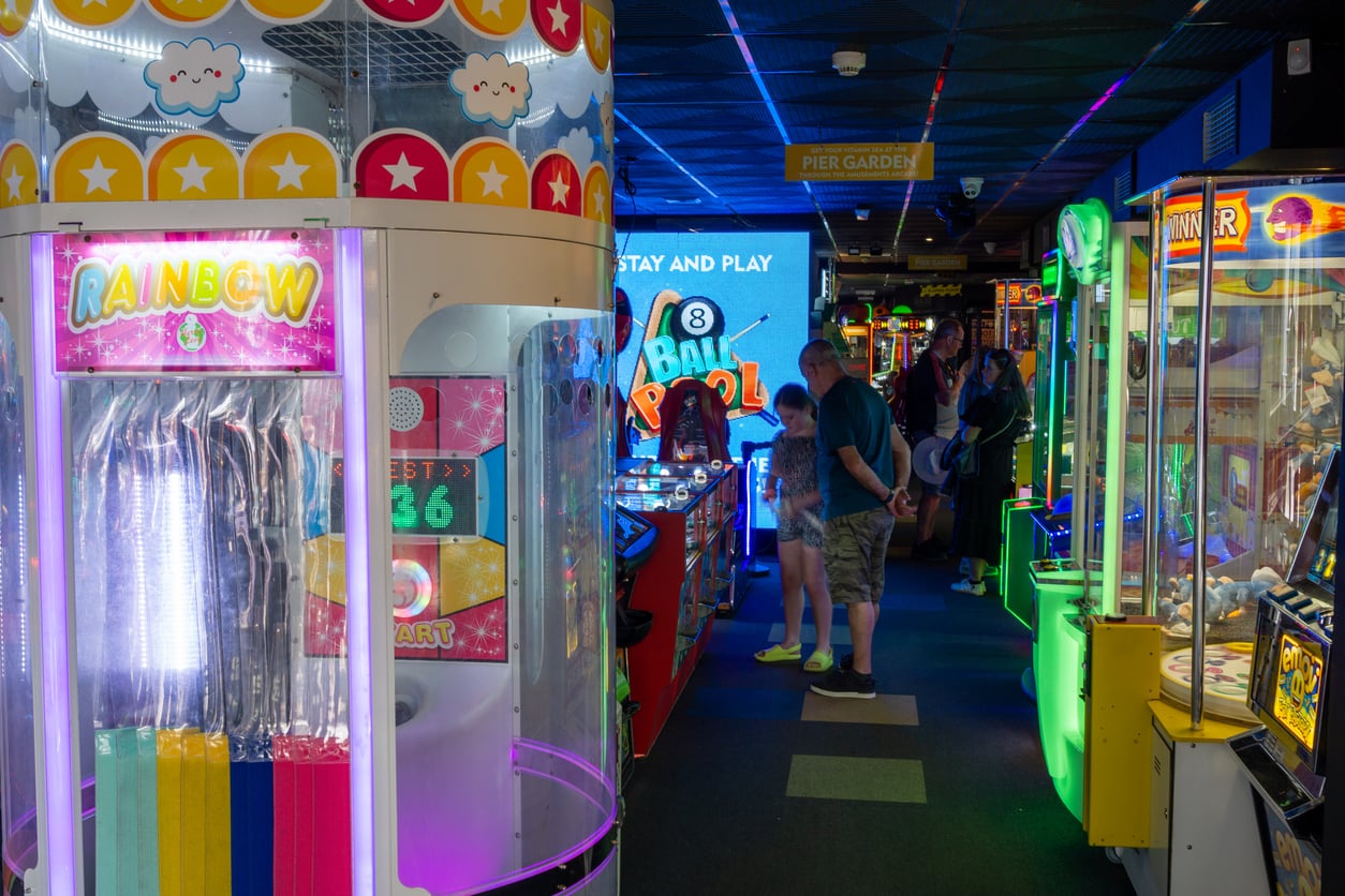 A brightly lit arcade filled with colorful machines and games, including a claw machine and a ticket dispenser, bustling with people enjoying the fun—and all set at nostalgic 1991 prices.