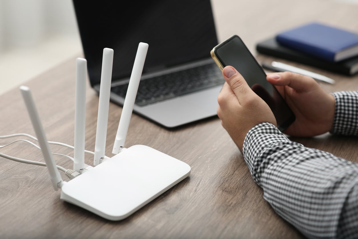 A person with DIY skills sits at a desk, holding a smartphone beside an open laptop, notebooks, and a white Wi-Fi router with four antennas.