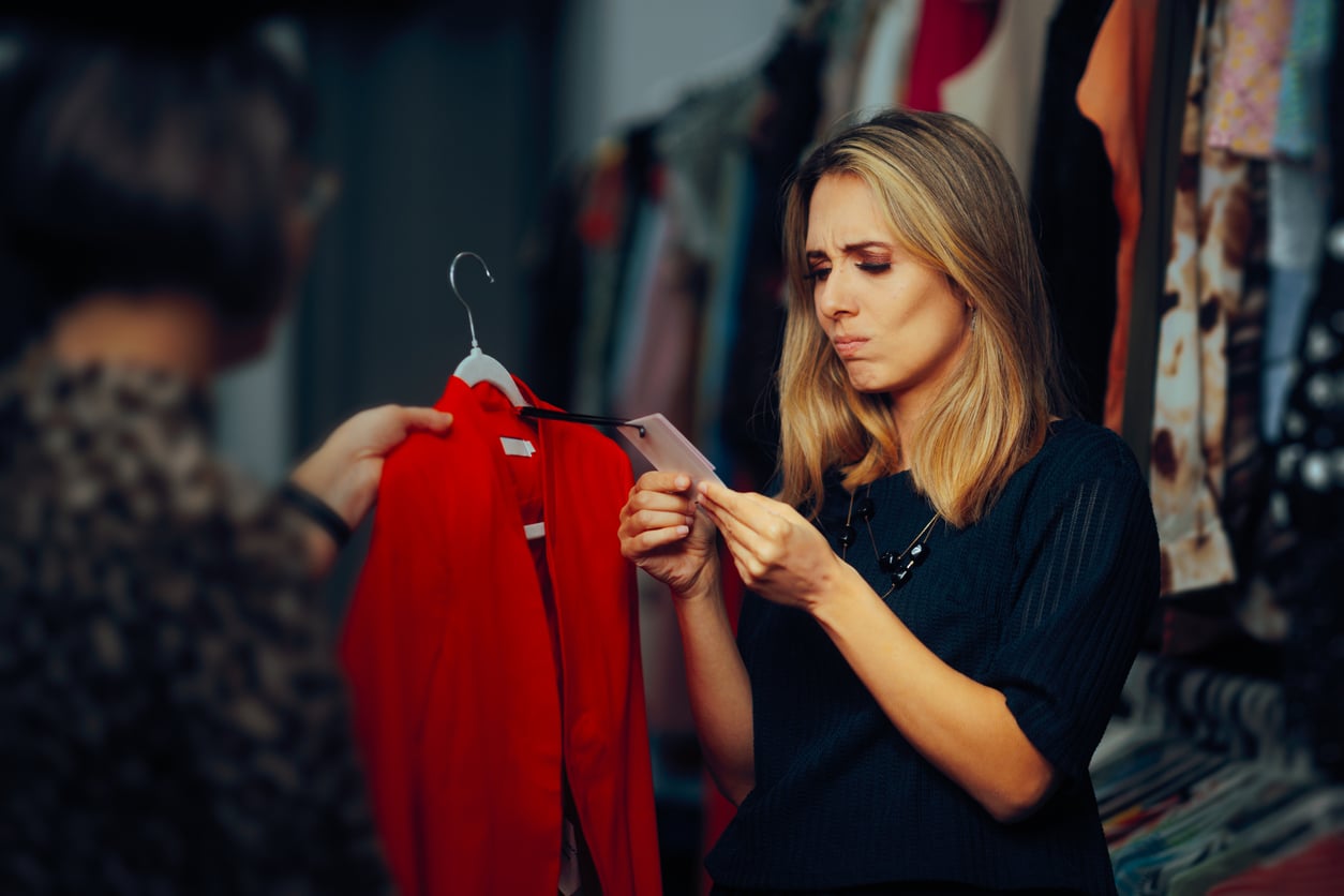 A woman in a clothing store looks intently at her phone, possibly weighing delayed purchases, while another person holds up a red jacket on a hanger. The woman appears unsure or concerned as dresses and clothes hang in the background.