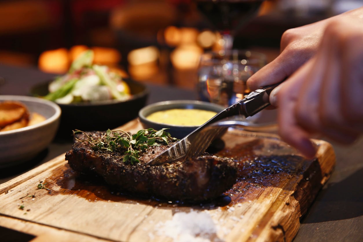 A hand cuts into a cooked steak garnished with herbs on a wooden board, with coarse salt nearby—a classic meal among products men still buy. Side dishes and a glass of wine are slightly blurred in the background.