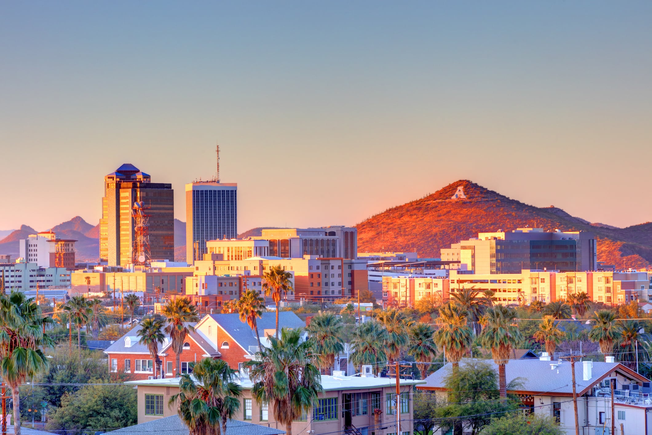Downtown Tucson, Arizona skyline at sunset with palm trees, modern and historic buildings, and a mountain in the background featuring a white "A" on its slope.